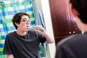 A teenage boy brushing his teeth with an electric tootbrush in front of the bathroom mirror.